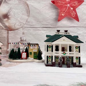 May include: A selection of miniature Christmas village houses, including a church, a shop, and two stately homes. The houses are white, red, and green, with intricate details. Two red star decorations and a large silver bauble are in the background.
