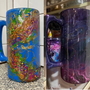May include: Two tall mugs with a unique galaxy-inspired design. One mug is blue with gold, green, and pink swirls and glitter. The other mug is purple with pink and white swirls and glitter.