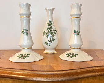 May include: Two cream-colored ceramic candle holders and a matching bud vase, each decorated with green holly leaves and red berries. The pieces feature gold trim accents on the rims and bases, with a smooth, glossy finish on the ceramic surfaces.