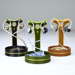 May include: Three in-ear monitor headphone stands in black, green, and brown. Each stand holds a pair of earphones with different designs and colours. The stands have a T-shape design with a circular base.