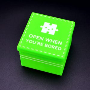 May include: A bright green square box with a white puzzle piece graphic and the text "OPEN WHEN YOU'RE BORED" in white. The box has a lid and is sitting on a dark surface. The box is a gift box.