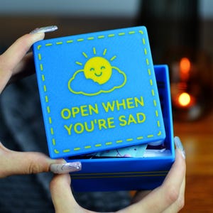 May include: A blue box with a yellow sun and cloud graphic, and the text "OPEN WHEN YOU'RE SAD". The box is open, revealing contents inside. The box has a stitched border design.