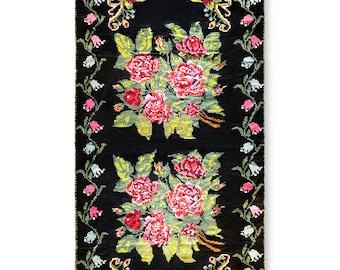 May include: A black rug with a floral design. The rug features pink, red, and green roses with green leaves. The border of the rug is a repeating pattern of white, pink, and green flowers.
