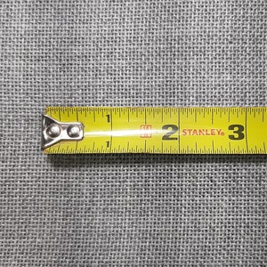 May include: A yellow Stanley tape measure with black markings, showing measurements in inches. The tape measure is partially extended, displaying the numbers 1, 2, and 3. The background is a gray, textured fabric.