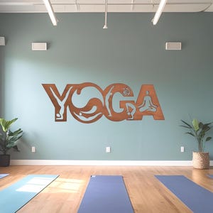 Puede incluir: Un letrero de pared met&aacute;lico de color marr&oacute;n con la palabra YOGA en letras may&uacute;sculas, donde cada letra contiene siluetas recortadas de personas realizando diversas posturas de yoga, montado en una pared azul claro sobre varias esterillas.