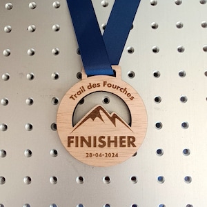 Puede incluir: Medalla de madera con una cinta azul. La medalla tiene una silueta de montaña y el texto "Trail des Fourches FINISHER 28-04-2024".