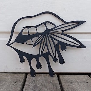 May include: Black metal wall art depicting a mouth with a cannabis leaf and dripping liquid. The artwork is set against a white wooden background and a wooden deck.