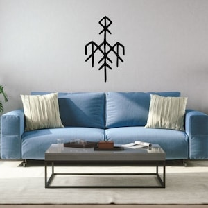 May include: A blue sofa with two pillows sits in front of a white wall with a black geometric design on it. A coffee table sits in front of the sofa with a book and a few glasses on it. A potted plant is on the left side of the image and a lamp is on the right side of the image.