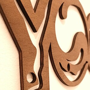 Puede incluir: Un recorte de pared de madera de color marr&oacute;n oscuro que presenta las letras Y y O en una fuente estilizada y conectada. El dise&ntilde;o incorpora la silueta de una persona en una postura de yoga integrada en la letra Y, con bordes lisos y acabado mate.