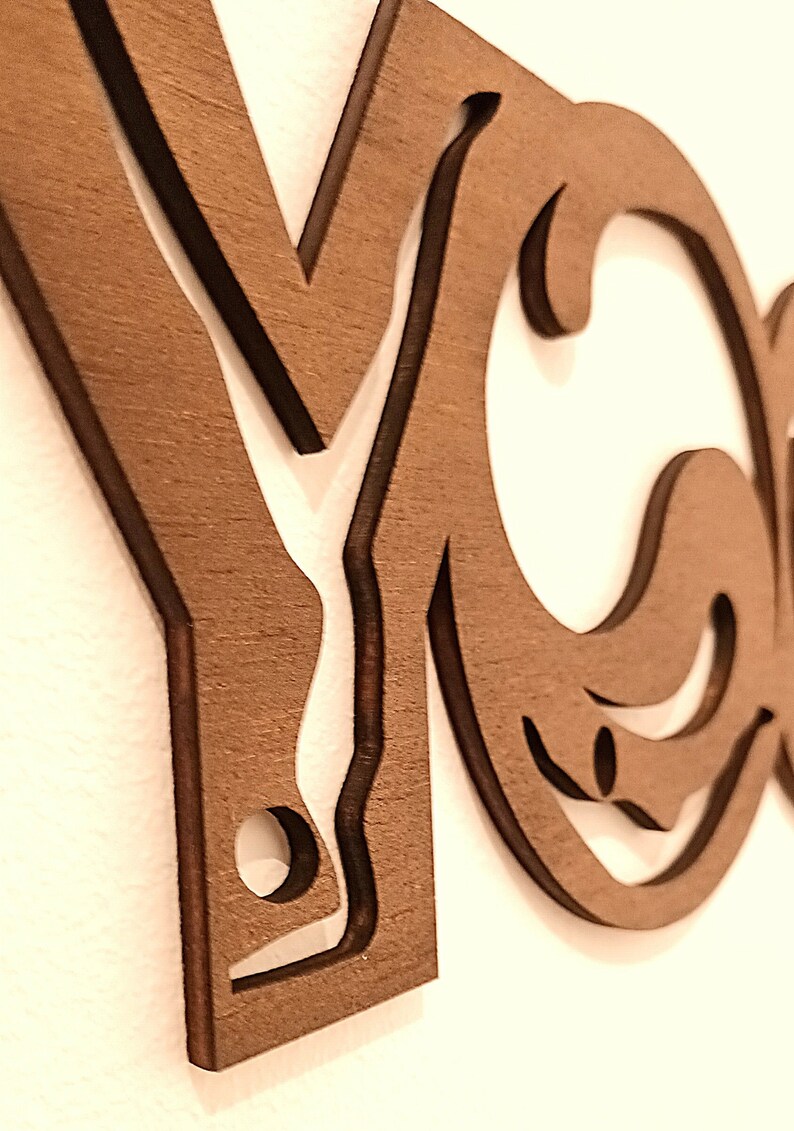 Puede incluir: Un recorte de pared de madera de color marr&oacute;n oscuro que presenta las letras Y y O en una fuente estilizada y conectada. El dise&ntilde;o incorpora la silueta de una persona en una postura de yoga integrada en la letra Y, con bordes lisos y acabado mate.