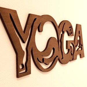 Puede incluir: Un recorte de pared de madera marr&oacute;n con la palabra YOGA en una fuente estilizada. Las letras incorporan siluetas de figuras en varias posturas, con la letra O conteniendo una forma circular y la letra A mostrando un s&iacute;mbolo Om.