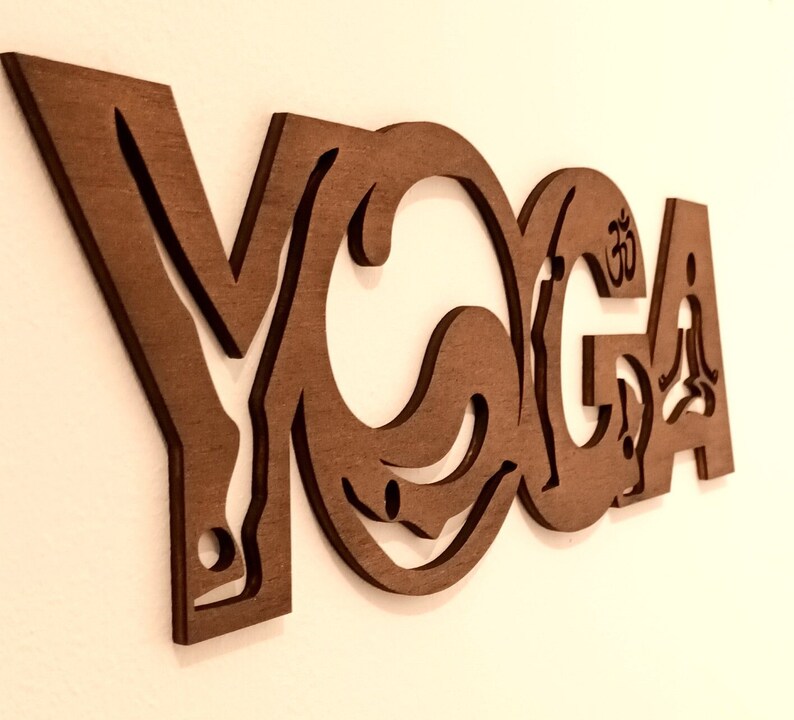 Puede incluir: Un recorte de pared de madera marr&oacute;n con la palabra YOGA en una fuente estilizada. Las letras incorporan siluetas de figuras en varias posturas, con la letra O conteniendo una forma circular y la letra A mostrando un s&iacute;mbolo Om.