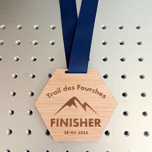 Op de afbeelding: Houten zeshoekige medaille met een blauw lint. De medaille heeft de tekst "Trail des Fourches" en "FINISHER" met de datum "28-04-2024" erop gegraveerd.