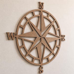 Puede incluir: Decoración de pared de madera con forma de rosa de los vientos en un tono marrón cálido. El diseño presenta una forma de estrella central con las letras direccionales N, E, S y W. La brújula es circular con un diseño abierto e intrincado.