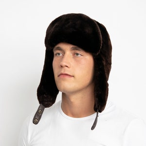 Pode incluir: Um chapéu trapper de pele sintética marrom com abas para as orelhas e uma tira de couro com dois botões. O chapéu está sendo usado por uma pessoa com uma camiseta branca.
