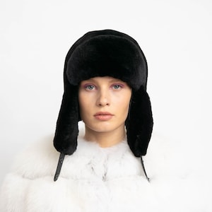 Könnte beinhalten: Ein schwarzer Kunstfell-Ushanka-Hut mit Ohrenklappen und Kinnriemen. Der Hut wird über einem weißen Pelzmantel getragen. Der Hut ist zum Schutz vor Kälte konzipiert.