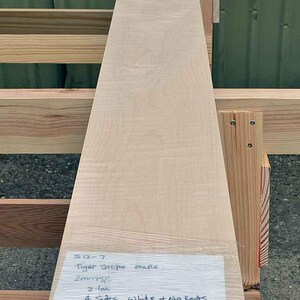 May include: A piece of tiger stripe maple wood, 20 inches by 175%, 2.1 metres long, with four sides white and no knots. The wood is AA figure. The wood is sitting on a wooden rack.