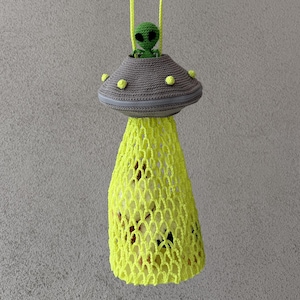 May include: A handmade crochet UFO-shaped bag with a green alien figure and a yellow net-like beam. The UFO is gray with yellow accents and a yellow strap. The text "Cow amigurumi is for display only is not included in the pattern" is visible.