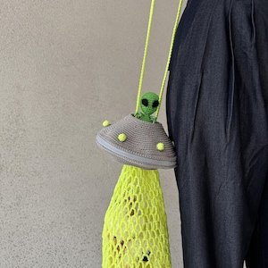 May include: A handmade crochet bag in the shape of a UFO, featuring a gray top with yellow accents and a neon yellow net bottom. The bag has a green alien figure inside. The text on the image says "Cow amigurumi is for display only is not included in the pattern."