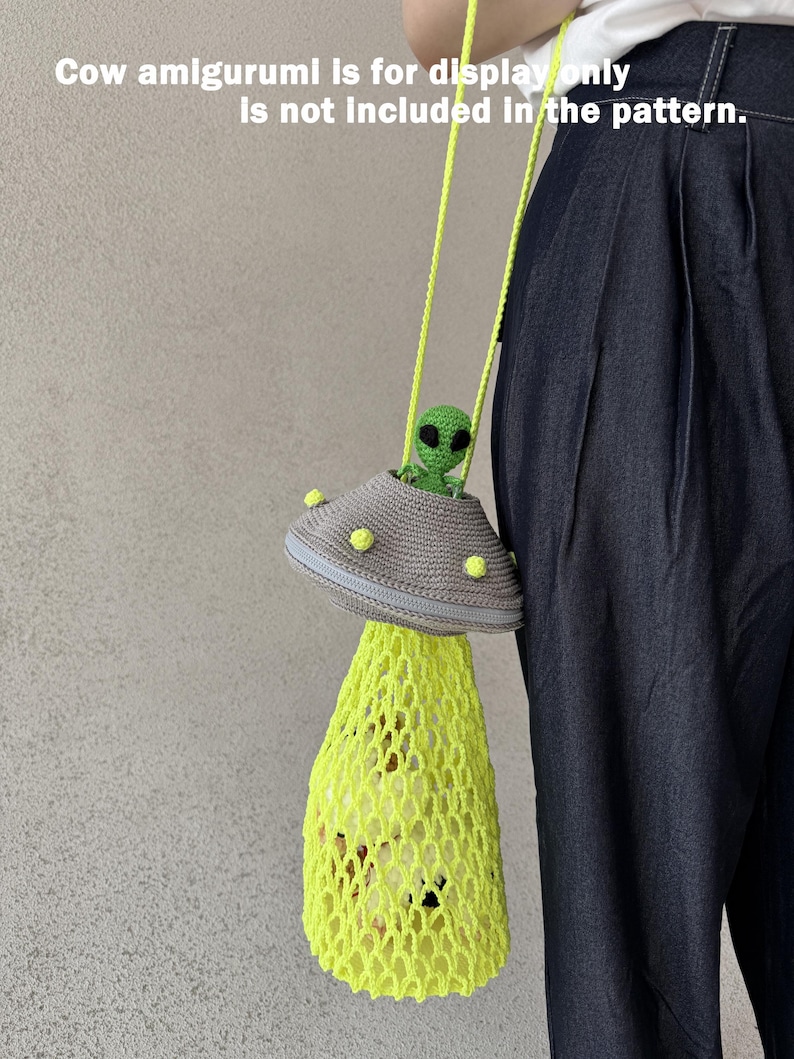 May include: A handmade crochet bag in the shape of a UFO, featuring a gray top with yellow accents and a neon yellow net bottom. The bag has a green alien figure inside. The text on the image says "Cow amigurumi is for display only is not included in the pattern."