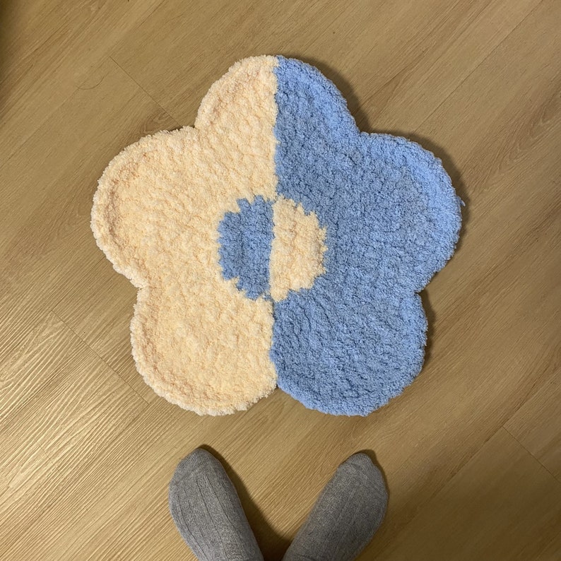 May include: A light blue and cream colored flower-shaped rug with a smaller flower design in the center. The rug is made of fluffy yarn.