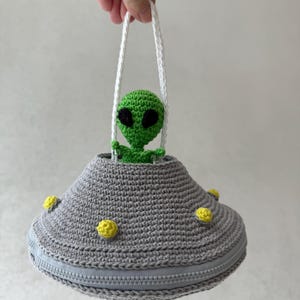 May include: A crocheted gray UFO-shaped purse with a zipper closure and yellow accents. A green alien figure with black eyes is inside the UFO, and the purse has white handles. The purse is handmade.