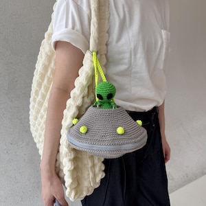 May include: A cream-colored, textured tote bag with a crocheted strap. A gray crocheted UFO-shaped purse with a green alien figure is attached. The purse has a blue zipper and yellow accents. The alien has black eyes.