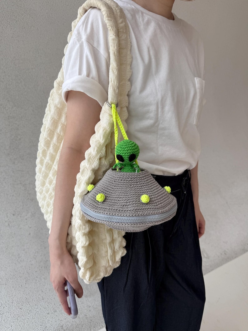 May include: A cream-colored, textured tote bag with a crocheted strap. A gray crocheted UFO-shaped purse with a green alien figure is attached. The purse has a blue zipper and yellow accents. The alien has black eyes.