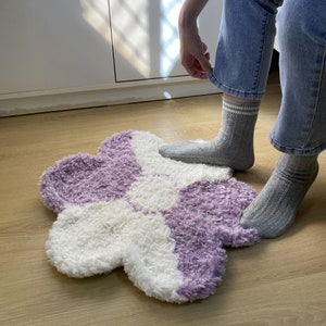 May include: A fluffy, two-toned rug in the shape of a flower. The rug is white and purple, with a soft, fuzzy texture.