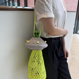 May include: A crocheted UFO-shaped bag with a green alien figure on top. The bag features a gray top with yellow accents and a neon yellow net-like bottom. The bag has a yellow strap and is being held by a person.