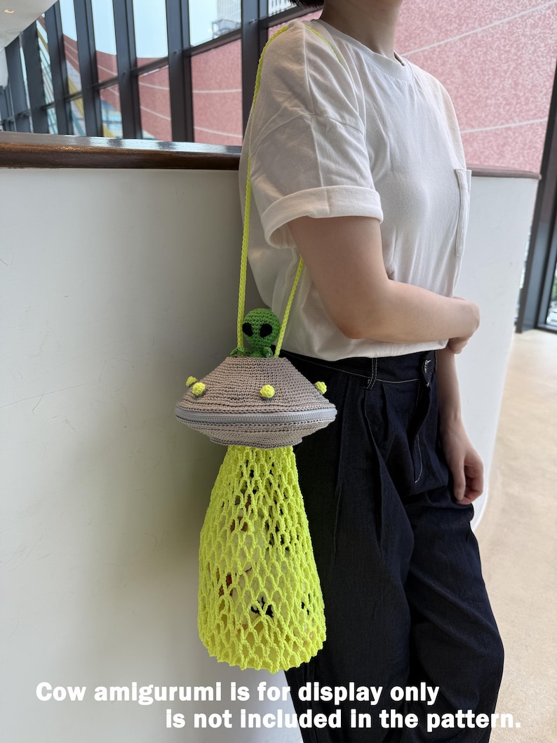 May include: A crocheted UFO-shaped bag with a green alien figure on top. The bag features a gray top with yellow accents and a neon yellow net-like bottom. The bag has a yellow strap and is being held by a person.