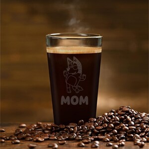 May include: A glass of dark coffee with a cartoon character etched on the front, with the word "MOM" below. Steam rises from the coffee. Coffee beans are scattered on the wooden surface.