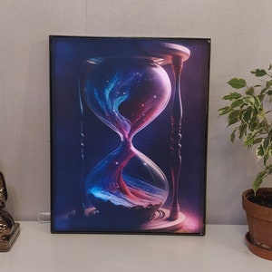 May include: A framed print of an hourglass with a galaxy inside. The hourglass is made of glass and has a wooden frame. The galaxy is a swirling mix of blue, pink, and purple.