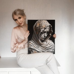 May include: A woman is holding a framed print of a sculpture depicting a woman's head and shoulders. The sculpture is made of white marble and has a gold-coloured veil over her head. The background of the print is black.