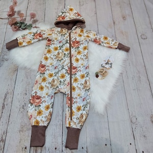 Könnte beinhalten: Ein weißer Baby-Strampler mit einem Blumendruck in Gelb-, Orange- und Brauntönen. Der Strampler hat eine braune Kapuze und einen Reißverschluss vorne.