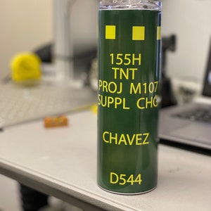 May include: A green tumbler with a yellow inscription that reads "155H TNT PROJ M107 SUPPL CHG CHAVEZ D544".
