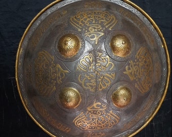 Puede incluir: Un escudo redondo de metal con un borde dorado y diseños dorados intrincados. El escudo tiene cinco acentos redondos dorados.