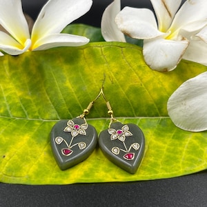 May include: Heart-shaped earrings with gold hooks. The earrings are a muted green colour and feature a floral design with red and white accents. The earrings are displayed on a green leaf with white flowers.