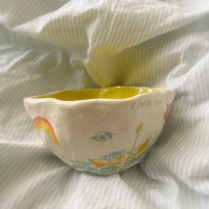 Op de afbeelding: Een witte keramische kom met een gele rand en handgeschilderde details. De kom heeft een regenboog, een oog en een kano met peddels. De tekst "Canoe Trip" staat aan de zijkant van de kom.