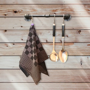 Op de afbeelding: Een bruin en zwart geruite keukendoek hangt aan een metalen pijpbeugel met twee zilveren kooklepels die aan haken hangen.