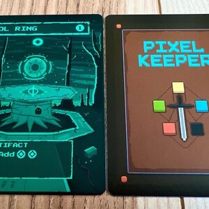 May include: A green and black card game called "Pixel Keeper" with a sword and colored squares on the front. The back of the card shows a green and black illustration of a ring with a glowing eye in the center.