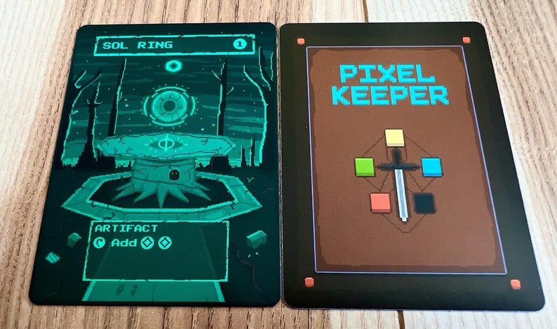 May include: A green and black card game called "Pixel Keeper" with a sword and colored squares on the front. The back of the card shows a green and black illustration of a ring with a glowing eye in the center.