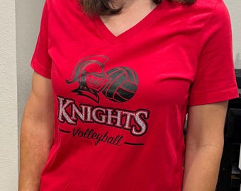 May include: A bright red short-sleeved V-neck t-shirt featuring a black graphic of a knight helmet and a volleyball. The word KNIGHTS is printed in large, bold, silver-glittered letters, with the word Volleyball written in black script below it.