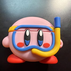 May include: A pink, spherical character case featuring a blue diving mask with a yellow strap and a yellow snorkel. The character has large black and blue eyes and red circular cheeks. The base of the case is red, and the overall design is smooth.
