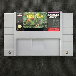 Peut inclure: Une cartouche de jeu vidéo grise Super Nintendo pour le jeu Secret of Mana. L'étiquette présente une illustration de forêt verte, le texte Secret of Mana, Square Soft et Super Nintendo Entertainment System, avec le code SNS-K2-USA visible.