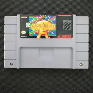 Könnte beinhalten: Eine graue Super Nintendo Entertainment System Spielkassette für EarthBound. Das Etikett zeigt eine goldene, stachelige Figur vor einem bunten, wirbelnden Hintergrund mit dem aufgedruckten Text EarthBound, Nintendo, KA-Einstufung und SNS-M8-USA.