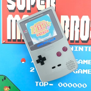 Puede incluir: Una consola de juegos portátil Game Boy Color plateada con una pantalla azul y blanca que muestra el juego "The Legend of Zelda: Oracle of Seasons". La consola tiene un pad direccional y botones negros, y el texto "GAME BOY COLOR" y "Nintendo" está impreso en la parte delantera.