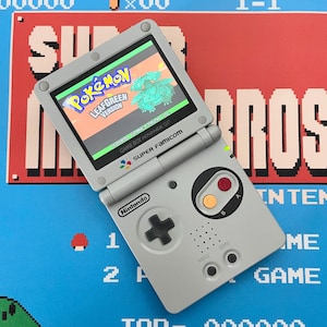 May include: A gray Nintendo Game Boy Advance SP console, open to display the "Pokémon LeafGreen Version" game. The console features a directional pad, A and B buttons, and a Super Famicom logo. The background has a pixelated video game theme.