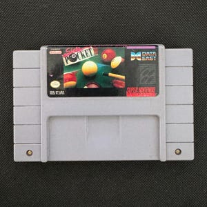 Op de afbeelding: Een grijze Super Nintendo videogame cartridge voor het spel Side Pocket. Het label toont een biljarttafel met kleurrijke biljartballen en een keu. Het Data East logo is zichtbaar in de rechterbovenhoek en het Nintendo zegel staat links.