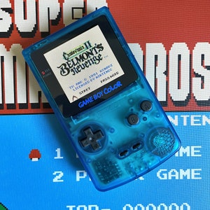 Puede incluir: Una consola de juegos portátil Game Boy Color azul con un cartucho de juego Castlevania II: Belmont's Revenge insertado. La pantalla muestra el título del juego y la información de copyright. La consola tiene un esquema de color azul y negro y presenta un pad direccional, botones y un botón de inicio.